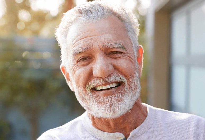 Smiling senior man with white beard in casual white shirt outdoors during daytime.