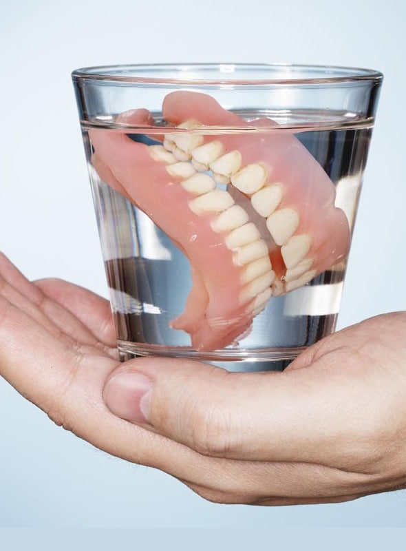Hand holding glass of water with dentures submerged inside for cleaning and storage.