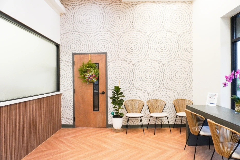 Modern dental waiting room with wooden door, chairs, plants, and whiteboard on wall.